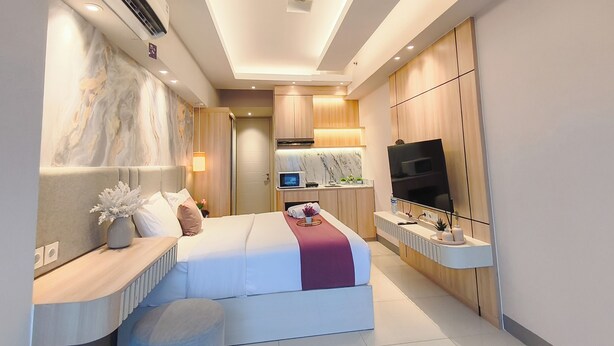 A modern hotel room with a bed, bedside table, TV, and built-in kitchen area. - Comfortable Studio Connected To Pakuwon Mall At Anderson Supermall Mansion Apartment (Surabaya)