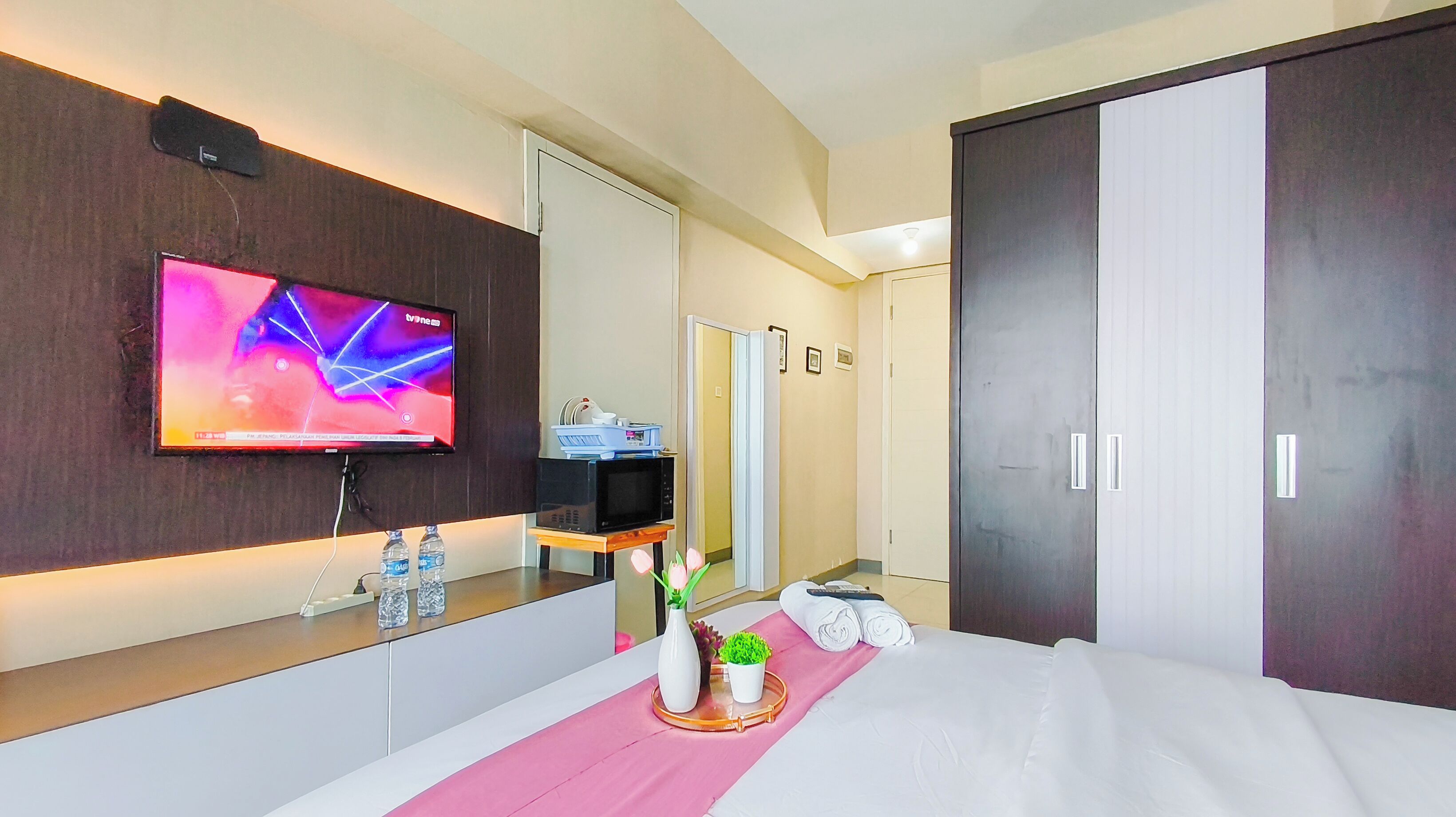 Habitación de hotel con televisor de pantalla plana, microondas, una mesita con botellas de agua, un jarrón con flores y una cama con ropa blanca.