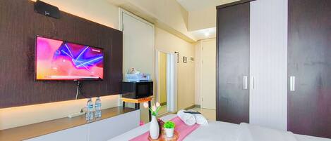 Habitación de hotel con televisor de pantalla plana, microondas, una mesita con botellas de agua, un jarrón con flores y una cama con ropa blanca.