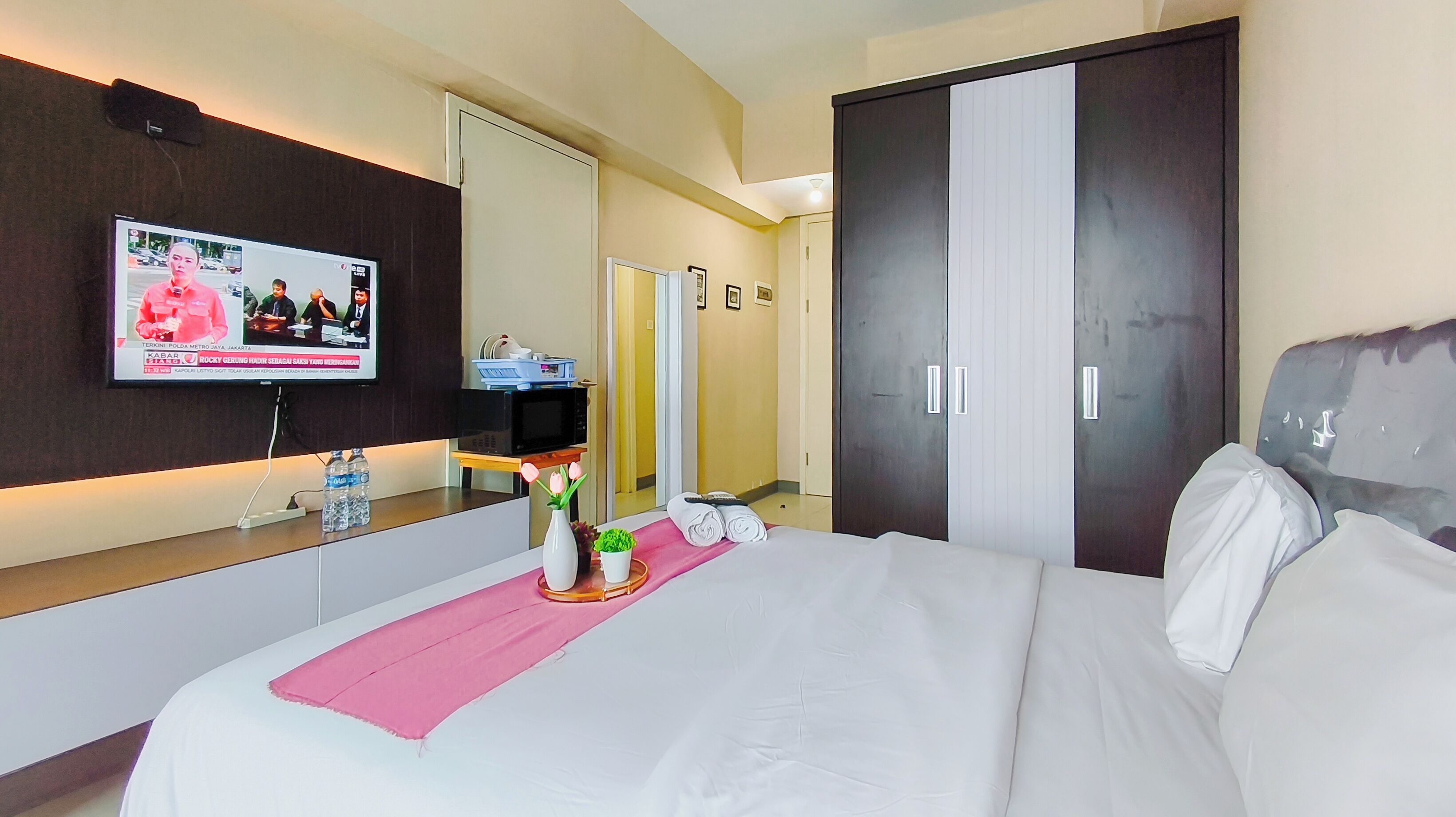 A hotel room with a bed, a TV displaying a news broadcast, a minibar, and a mirror.