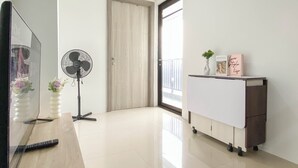 A room with a TV, a standing fan, a white cabinet, and a door leading to a balcony. - Contemporary And Restful 2Br At Apartment Anwa Residence (South Tangerang)