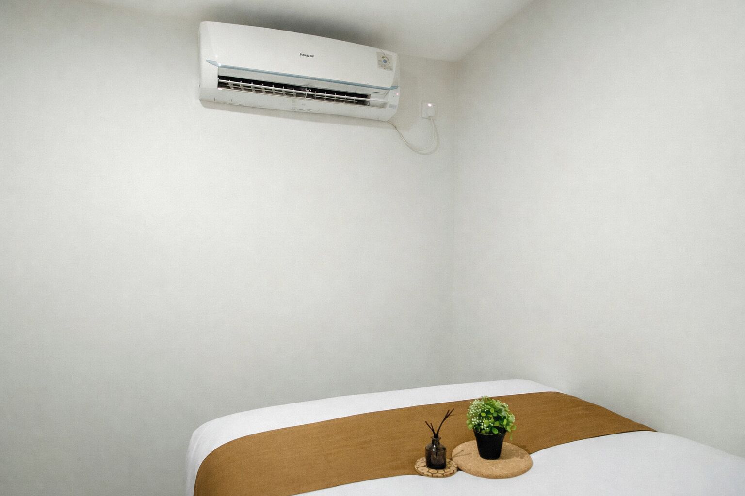 Una habitación con una cama, una mesita con una planta y un jarrón, y un equipo de aire acondicionado en la pared.