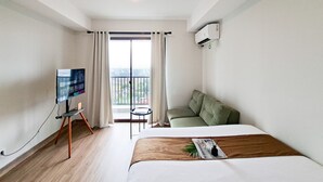 A hotel room with a bed, a television on a stand, a sofa, a balcony with a view, and an air conditioning unit. - Homey And Minimalist Studio Apartment At Anwa Residence (South Tangerang)