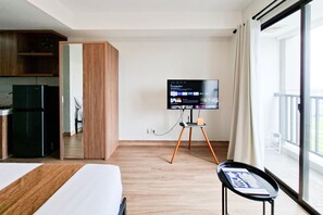 Une chambre d’hôtel avec un lit, un téléviseur posé sur un meuble, une petite table, un coin cuisine et un balcon avec des rideaux.