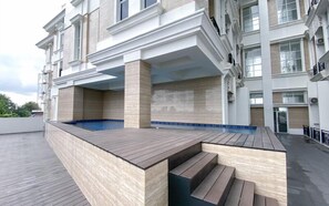 Area piscina sul tetto con scalini in legno che conducono alla vasca, circondata da un edificio moderno con numerosi balconi.
