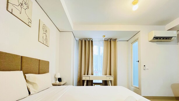 A hotel room with a bed, a desk, a chair, a window with curtains, and an air conditioning unit. - Studio Apartment Near Upn Veteran Yogyakarta At Barsa City (Depok)