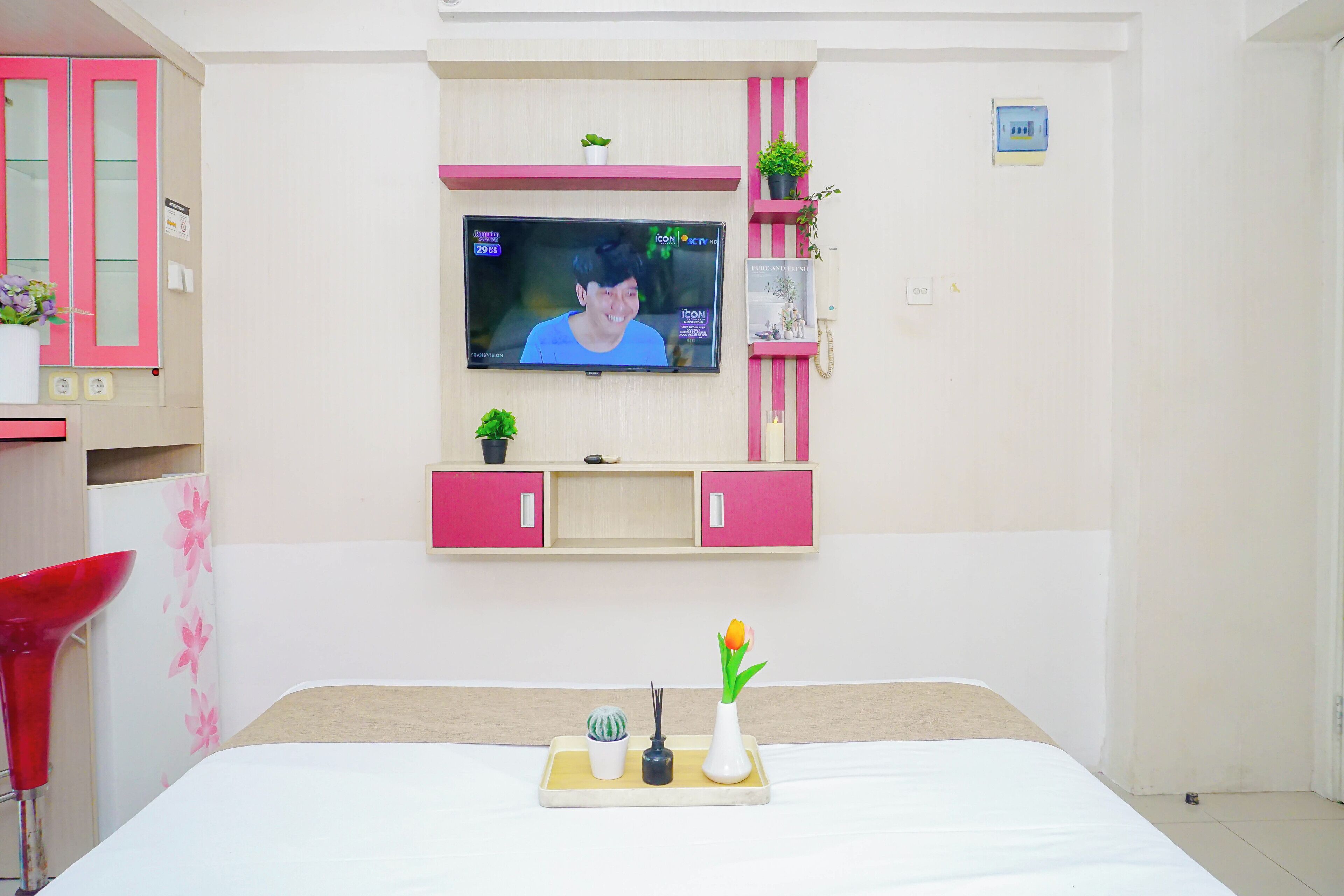 Een slaapkamer met een bed, een aan de muur bevestigde televisie, een klein plankje met planten en een dienblad met een vaas en een kaars.