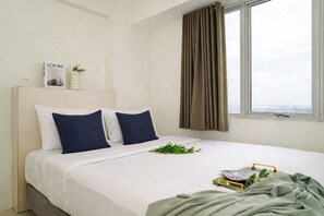 A neatly made bed with white and dark blue pillows, a small plant, and a green blanket on a white mattress. - Neat And Nice 2Br At Apartment Bassura City (Jakarta)