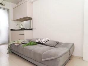 A compact room with a sofa bed, a small kitchenette with a sink and stove, and a white wall. - Neat And Nice 2Br At Apartment Bassura City (Jakarta)