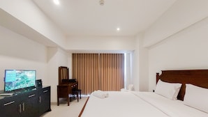 A hotel room with a large bed, a television on a dresser, a desk, and a window with curtains. - Comfy And Nice Studio Apartment At Carstensz Residence (Gading Serpong)