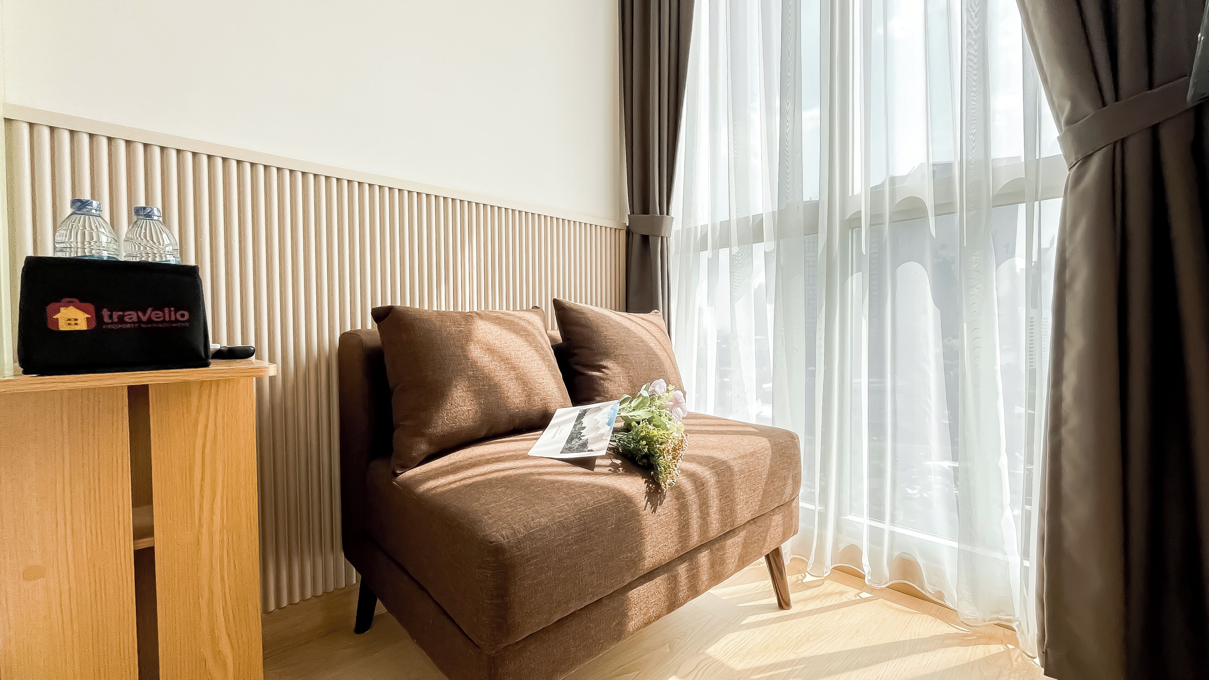 Ein braunes Sofa mit einem Kissen und einem Blumenstrauß, ein Holztisch mit einer schwarzen Tasche und zwei Wasserflaschen, sowie ein Fenster mit durchsichtigen Vorhängen.