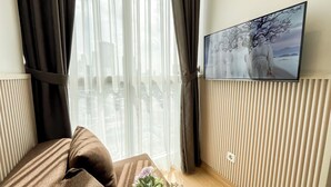 Ett rum med en säng, en tv som är monterad på väggen och ett fönster med tunna, genomskinliga gardiner.