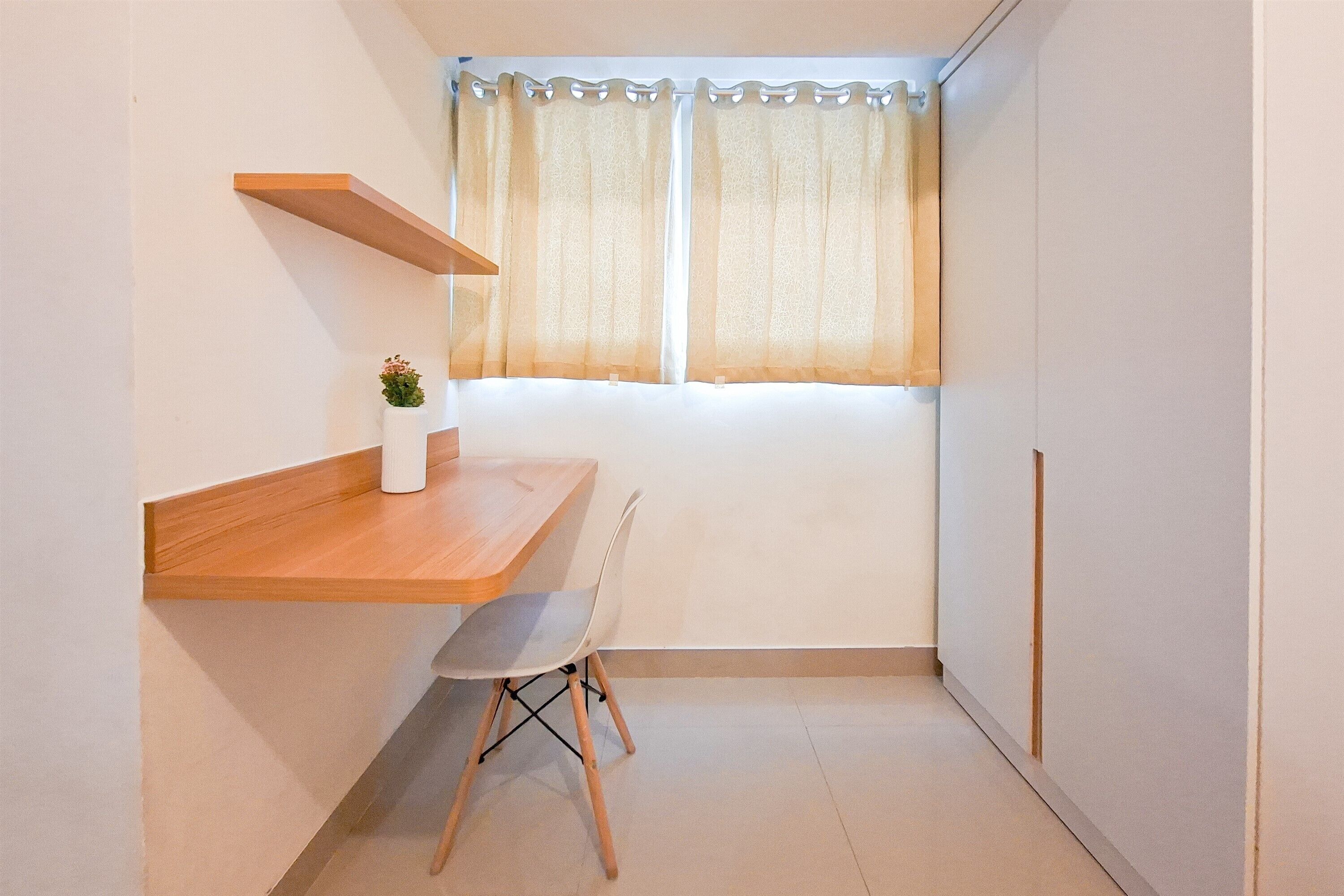 Een lichte, compacte kamer met een houten bureau, een stoel, een plank met een plant in pot en een raam met gordijnen.