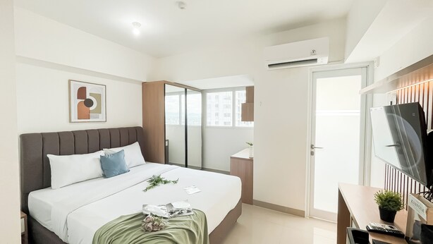 A hotel room with a bed, a desk with a computer, a mirror, and a window with a city view. - Homey And Minimalist Studio At Apartment Cisauk Point (Cisauk)