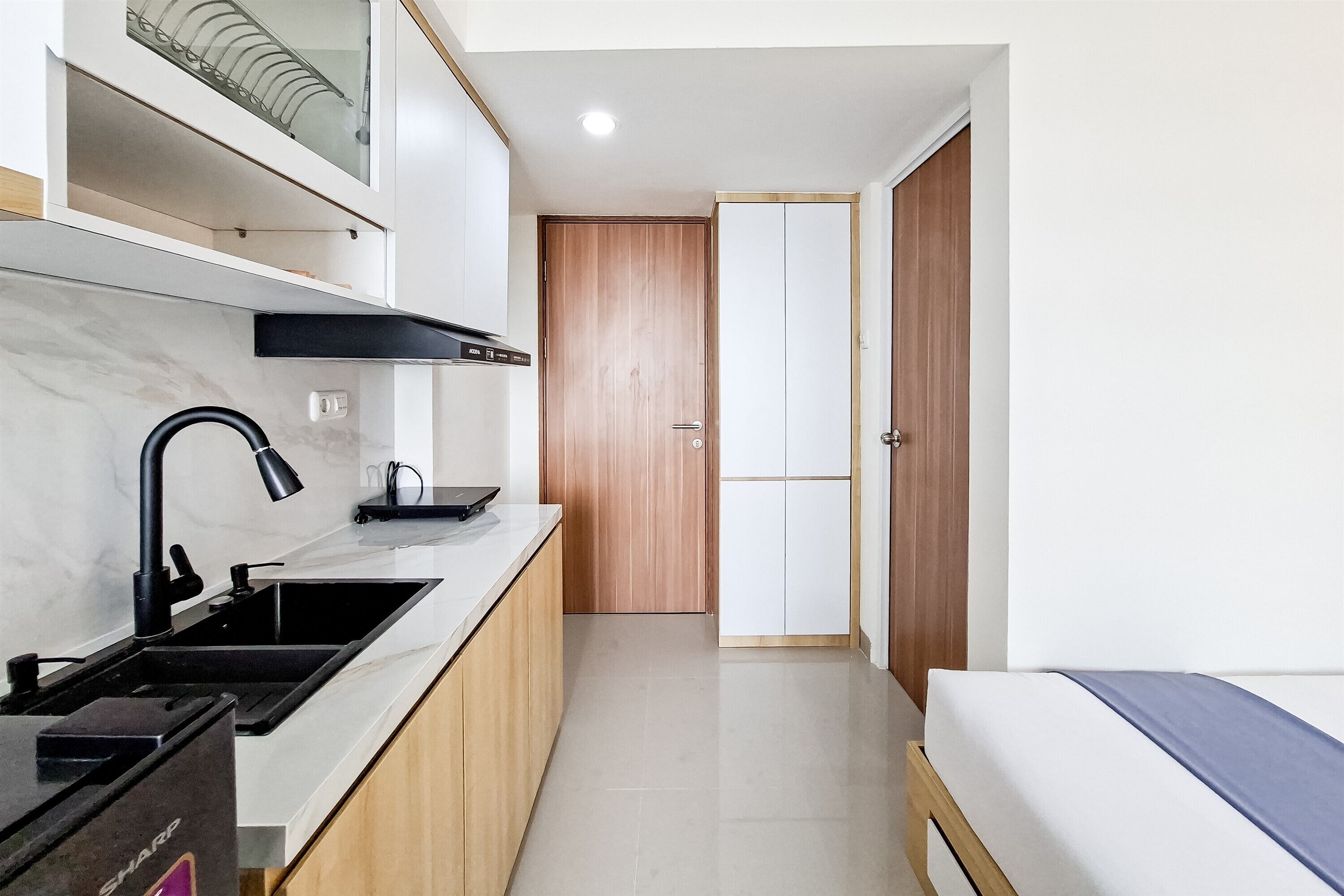 A narrow hallway with a kitchen area on one side, a wooden door, and a bed with a blue bedspread on the other.