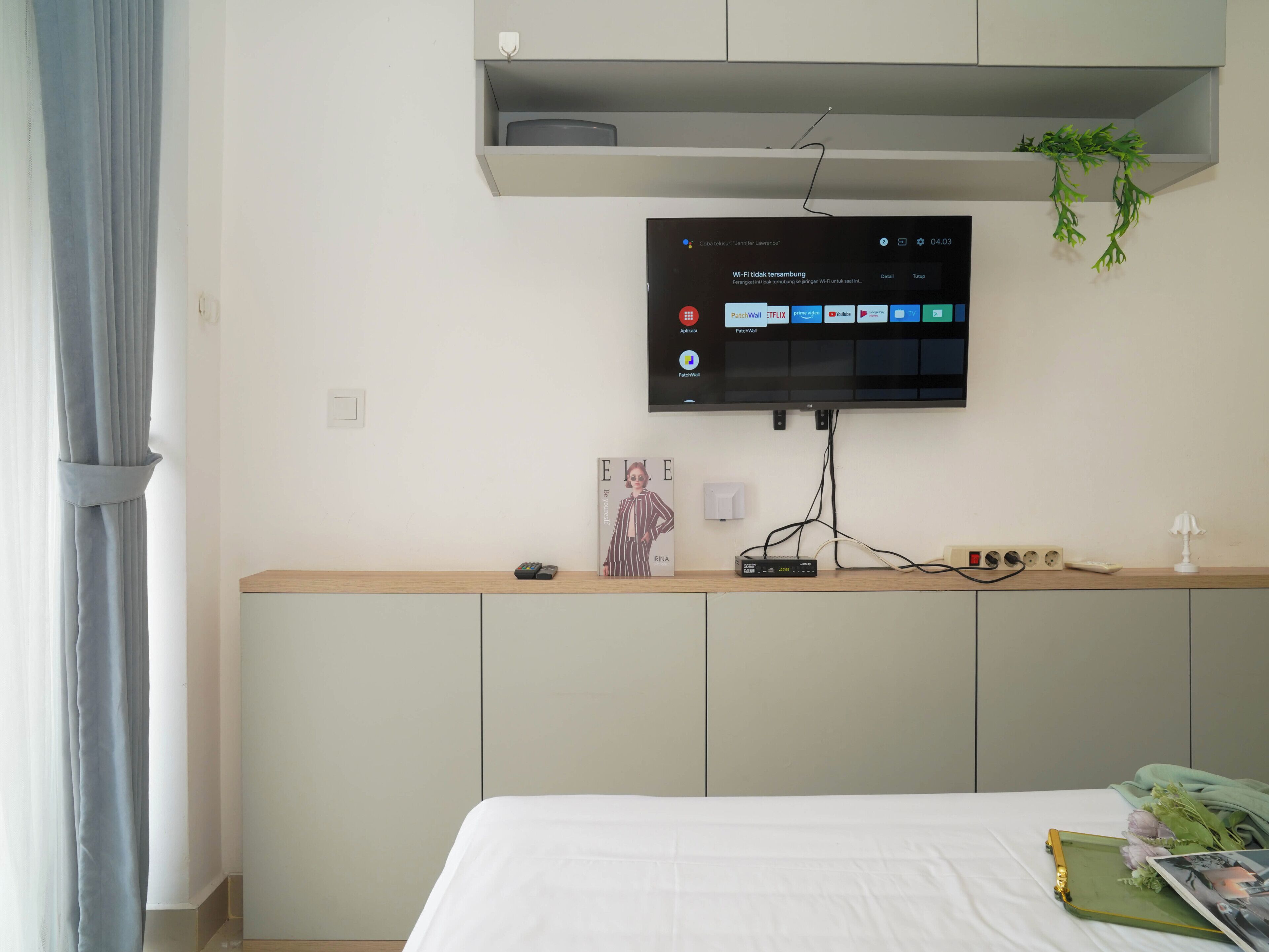 A bedroom with a bed, a TV mounted on the wall, a bedside table with a remote control, and a power strip.