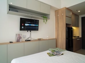 A modern hotel room with a bed, a flat-screen TV mounted on the wall, a small table with a remote control, and a refrigerator in the kitchen area. - Comfortable Space And Neat Studio At Apartment Citra Living (Jakarta)