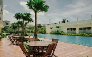 A poolside area with wooden tables and chairs, surrounded by palm trees and a swimming pool. - Comfortable Space And Neat Studio At Apartment Citra Living (Jakarta)
