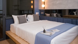A modern hotel room with a large bed, a bedside table, a mirror, and wall-mounted lights. - Deluxe 1Br Apartment Collins Boulevard (Tangerang)