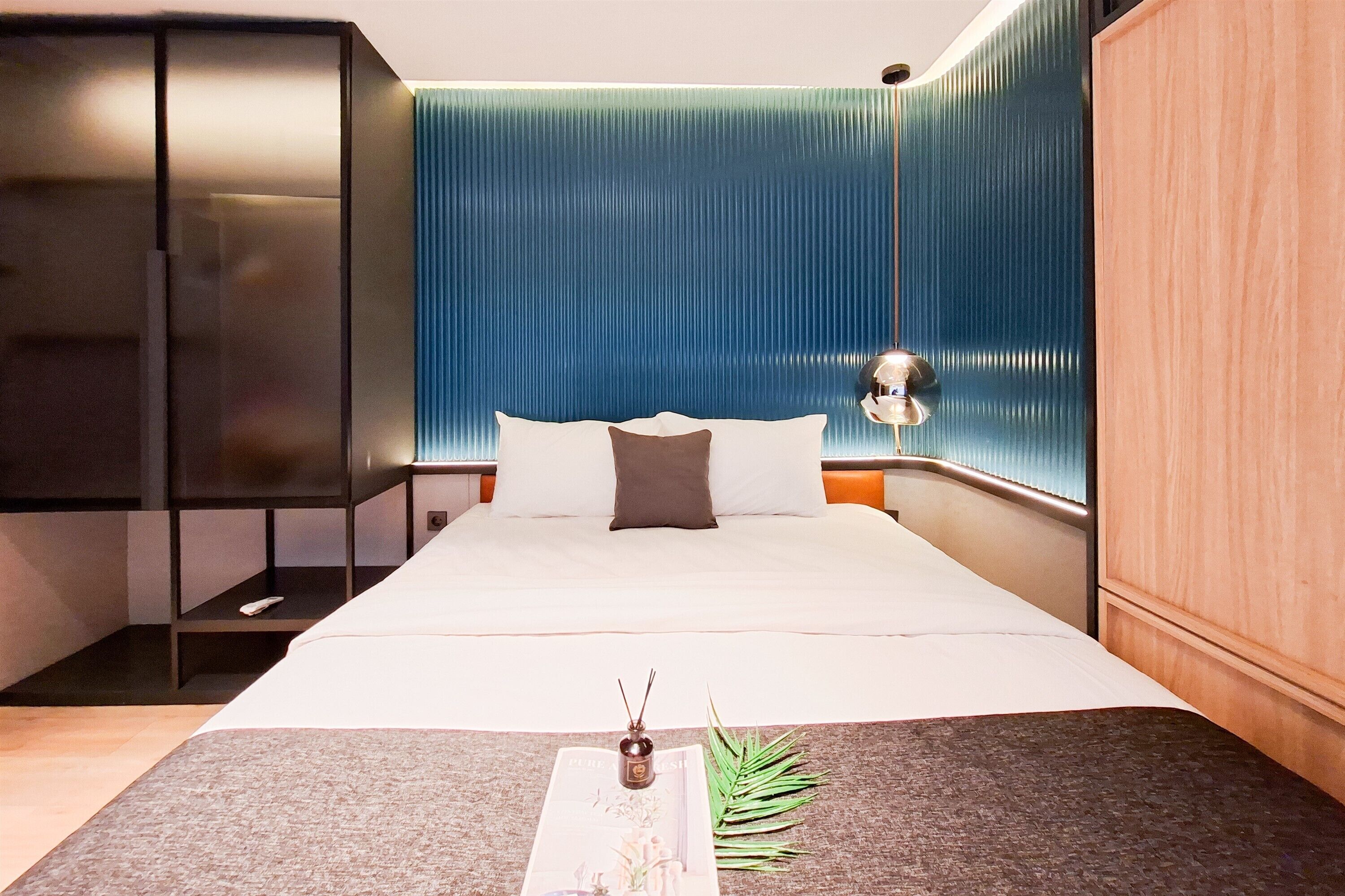 A modern hotel room with a large bed, a bedside table, a small plant, and a wall-mounted light fixture.