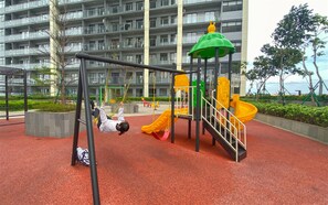 A playground with swings and slides, surrounded by a red surface and a multi-story building in the background. - Functional And Strategic Studio At Apartment Daan Mogot City (Jakarta)
