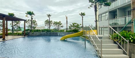 Une piscine avec un toboggan jaune, entourée de palmiers et dotée d’un espace abrité.