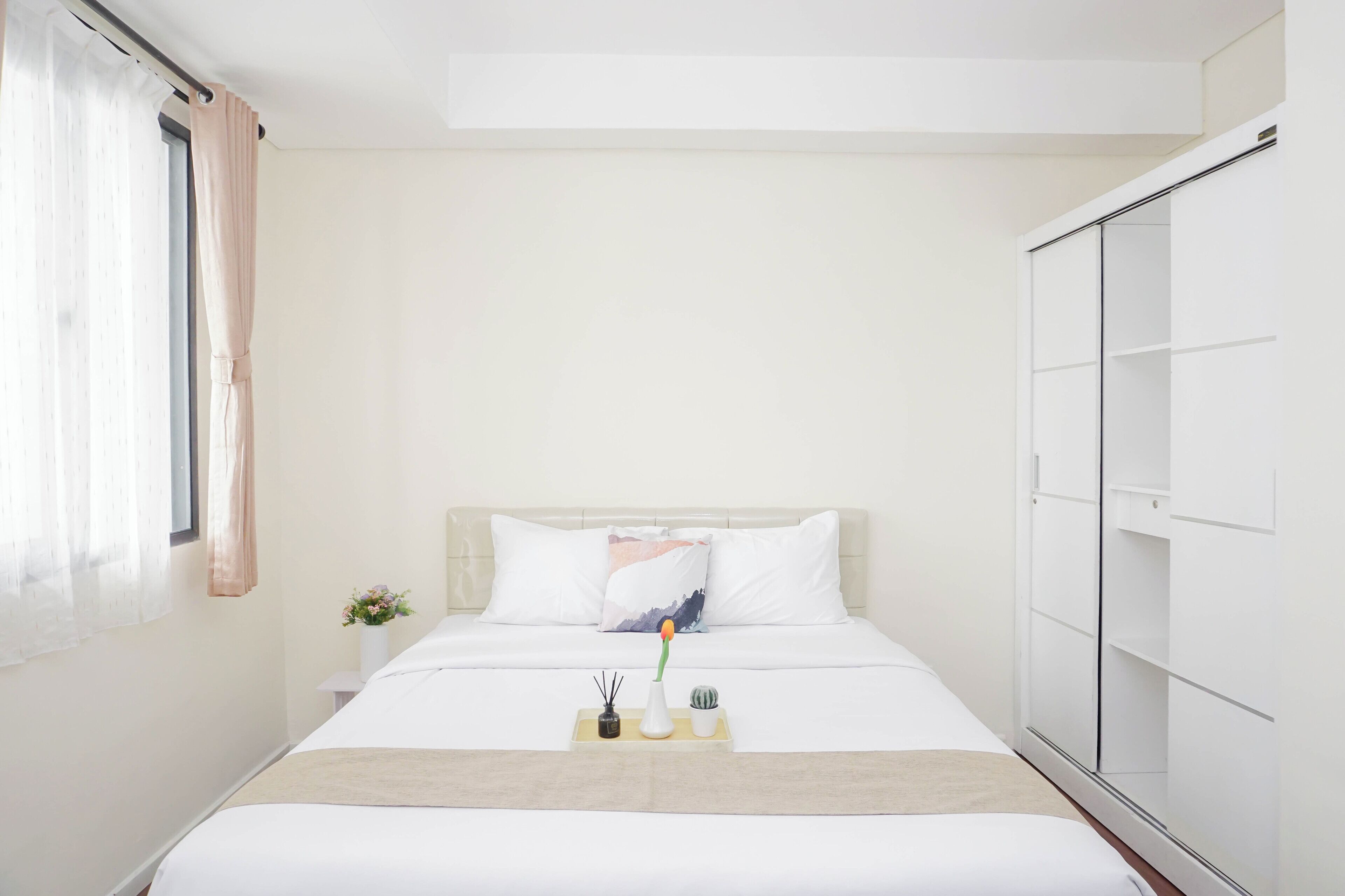Una habitación de hotel con una cama, una mesita de noche, un florero con flores, una planta pequeña y un armario.