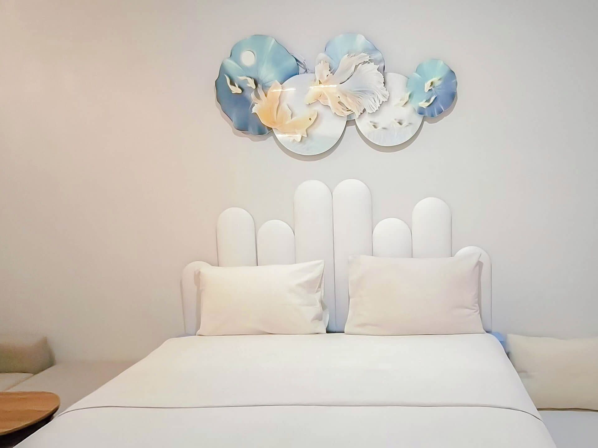 Een bed met een wit hoofdeinde en twee kussens, een muur met een decoratieve blauw-witte bloemversiering, en een houten nachtkastje aan de linkerkant.