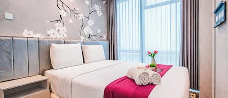 Habitación de hotel con cama, mesita de noche, pared decorada con motivos florales, ventana con cortinas y una pequeña planta sobre la cama.