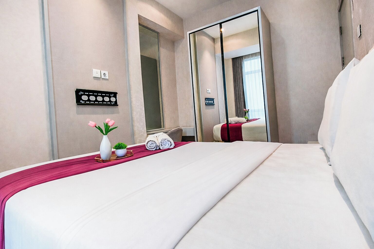 Habitación de hotel con una cama grande, un espejo, una mesita con un jarrón y una planta, y un panel de control en la pared.