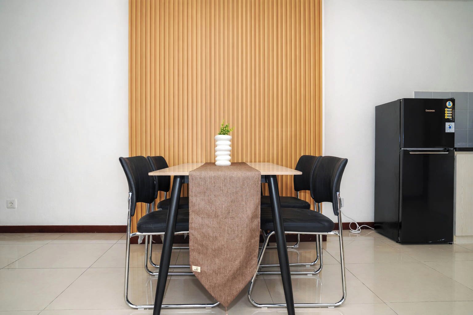 Un comedor con una mesa, cuatro sillas, una pequeña planta en maceta, un refrigerador y un panel de pared de madera.
