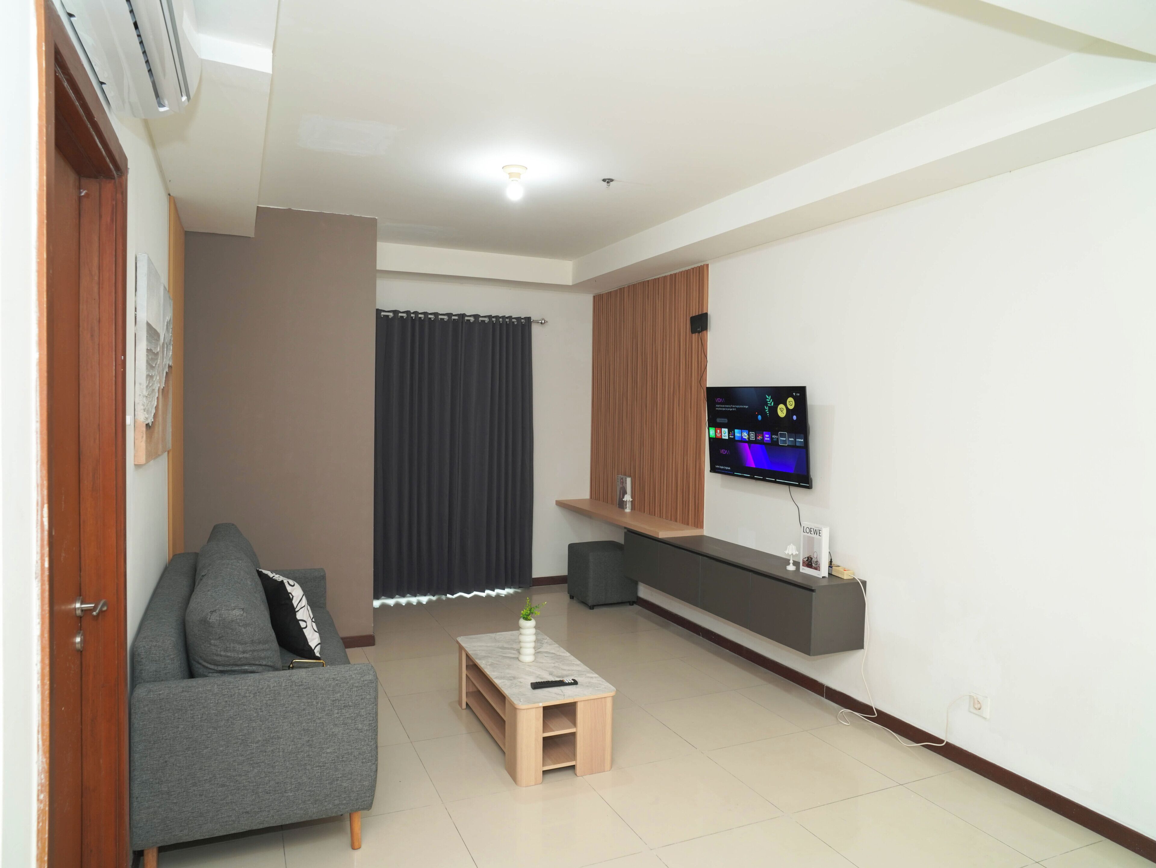 A living room with a grey sofa, a small wooden coffee table, a TV on a wall, and a curtain.