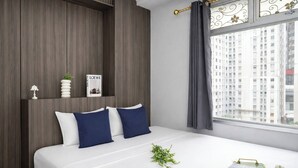 Una habitación de hotel con una cama, una ventana con cortinas, un estante con un libro y una planta pequeña.
