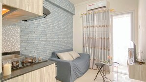 A compact living space with a kitchenette, a sofa, a small table, and a curtain with a floral pattern. - Warm And Restful 2Br At Green Pramuka City Apartment (Jakarta)