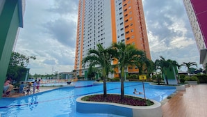 A swimming pool surrounded by a landscaped area with palm trees, a tall residential building in the background, and a clear sky. - Warm And Restful 2Br At Green Pramuka City Apartment (Jakarta)