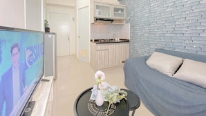 A modern living room with a flat-screen TV, a sofa, a small table with a vase, and a kitchen area with a brick wall and cabinets. - Warm And Restful 2Br At Green Pramuka City Apartment (Jakarta)