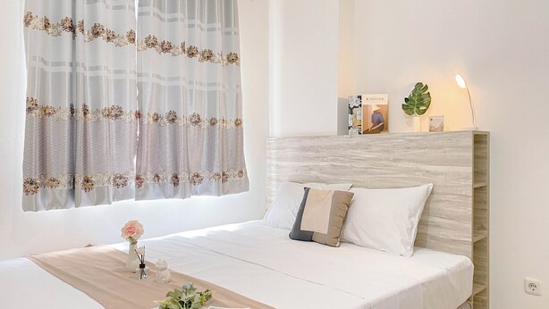 A neatly made bed with a beige bedspread, a white pillow, and a decorative vase with a flower. - Warm And Restful 2Br At Green Pramuka City Apartment (Jakarta)
