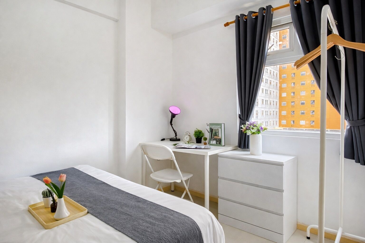 A bedroom with a bed, a desk, a chair, a bedside table, a window with curtains, and a view of buildings.