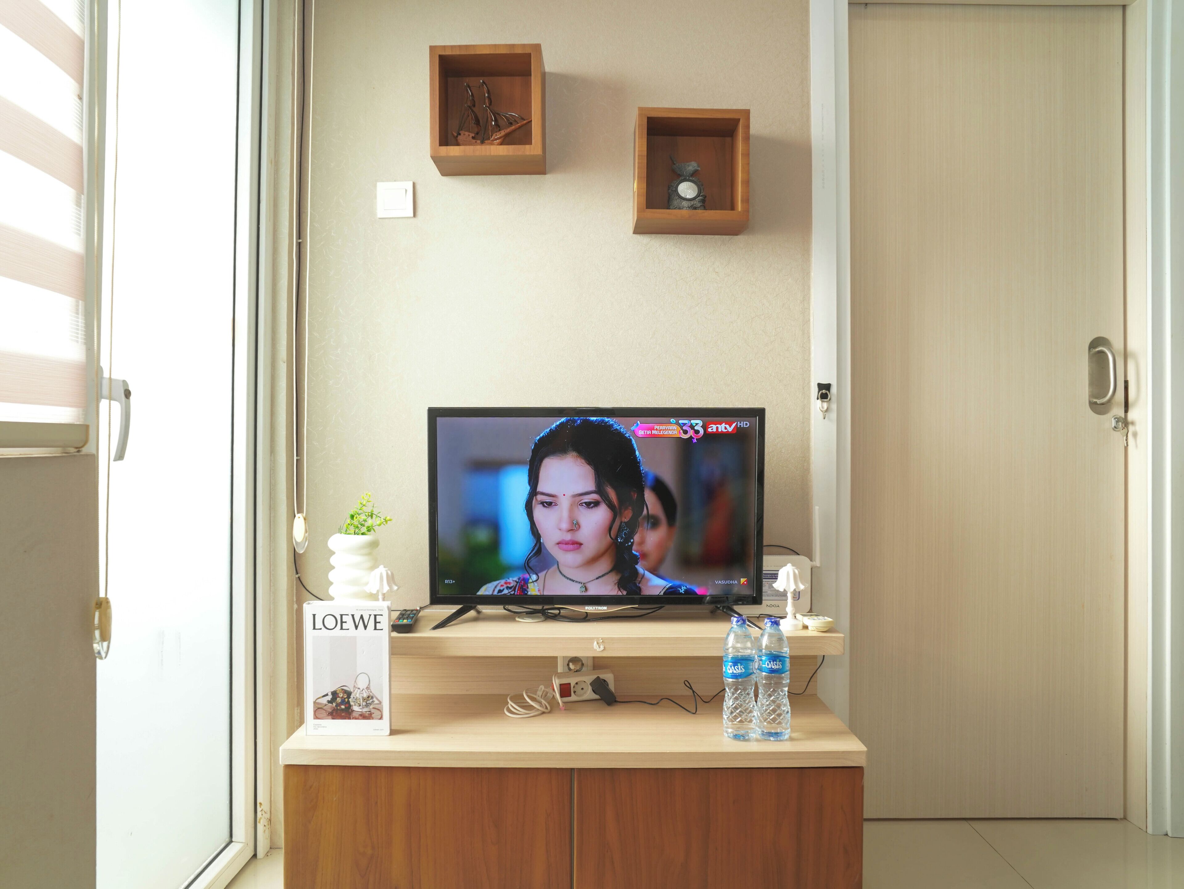 A television on a wooden stand with a remote, a small plant, and two water bottles on a table.