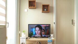 Pöydällä on televisio, joka on sijoitettu puiseen jalustaan, kaukosäädin, pieni kasvi ja kaksi vesipulloa.