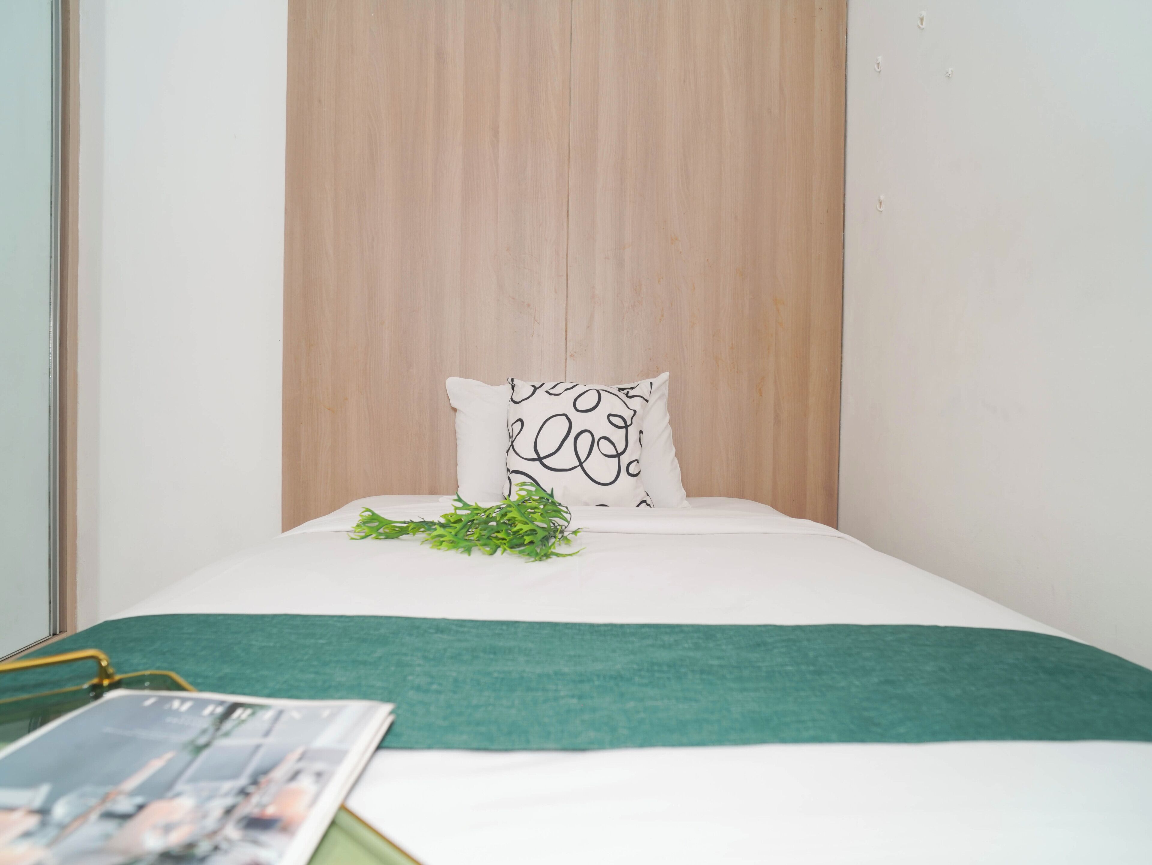A neatly made bed with a white and green bedspread, a single decorative pillow with a black pattern, and a small green plant on the bed.