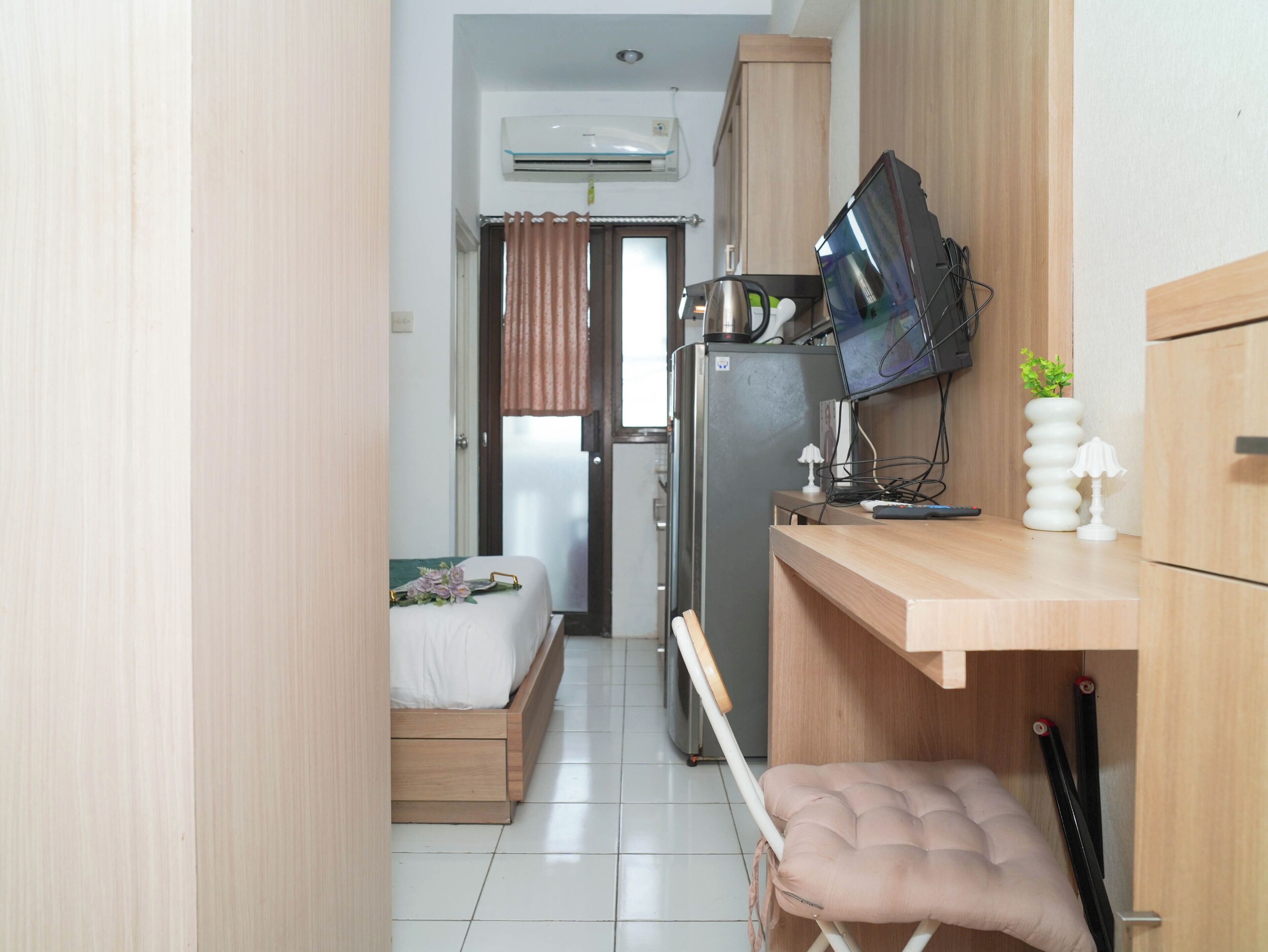 Een kleine hotelkamer met een bed, bureau, televisie, koelkast en een raam met gordijnen.