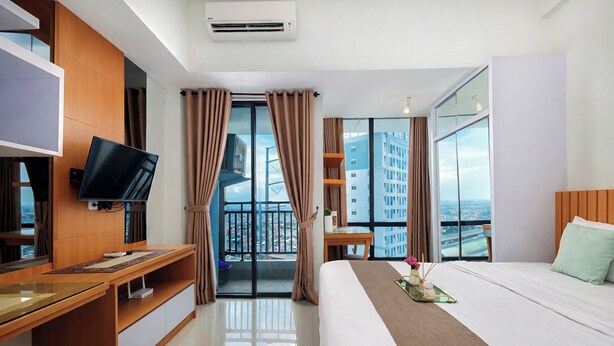 A hotel room with a bed, a desk with a flat-screen TV, a balcony with curtains, and a city view. - Urban Look Studio Apartment At Lrt City Bekasi (Bekasi)