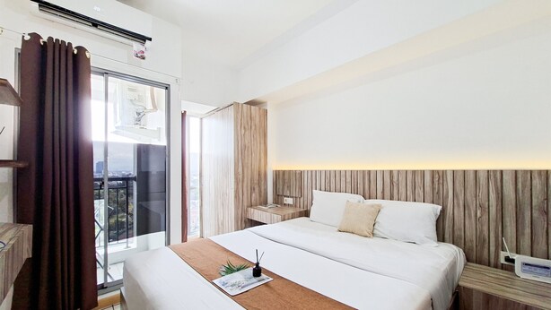 A hotel room with a large bed, a bedside table, a nightstand, a window with curtains, and a view of the outside. - Great Deal Studio Apartment At M-Town Residence (Gading Serpong)
