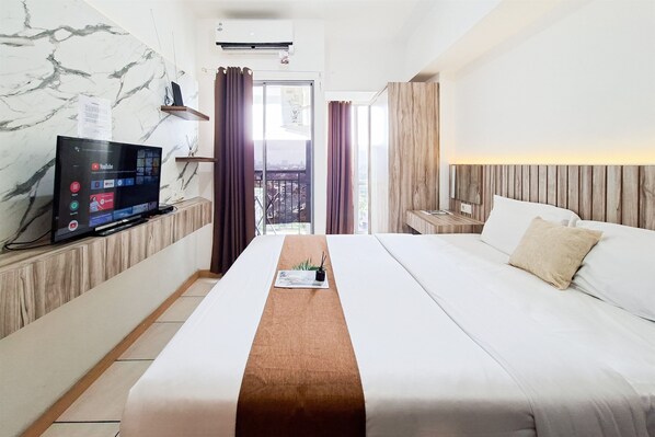 A hotel room with a bed, a TV, a wooden headboard, a view of the city, and a balcony. - Great Deal Studio Apartment At M-Town Residence (Gading Serpong)