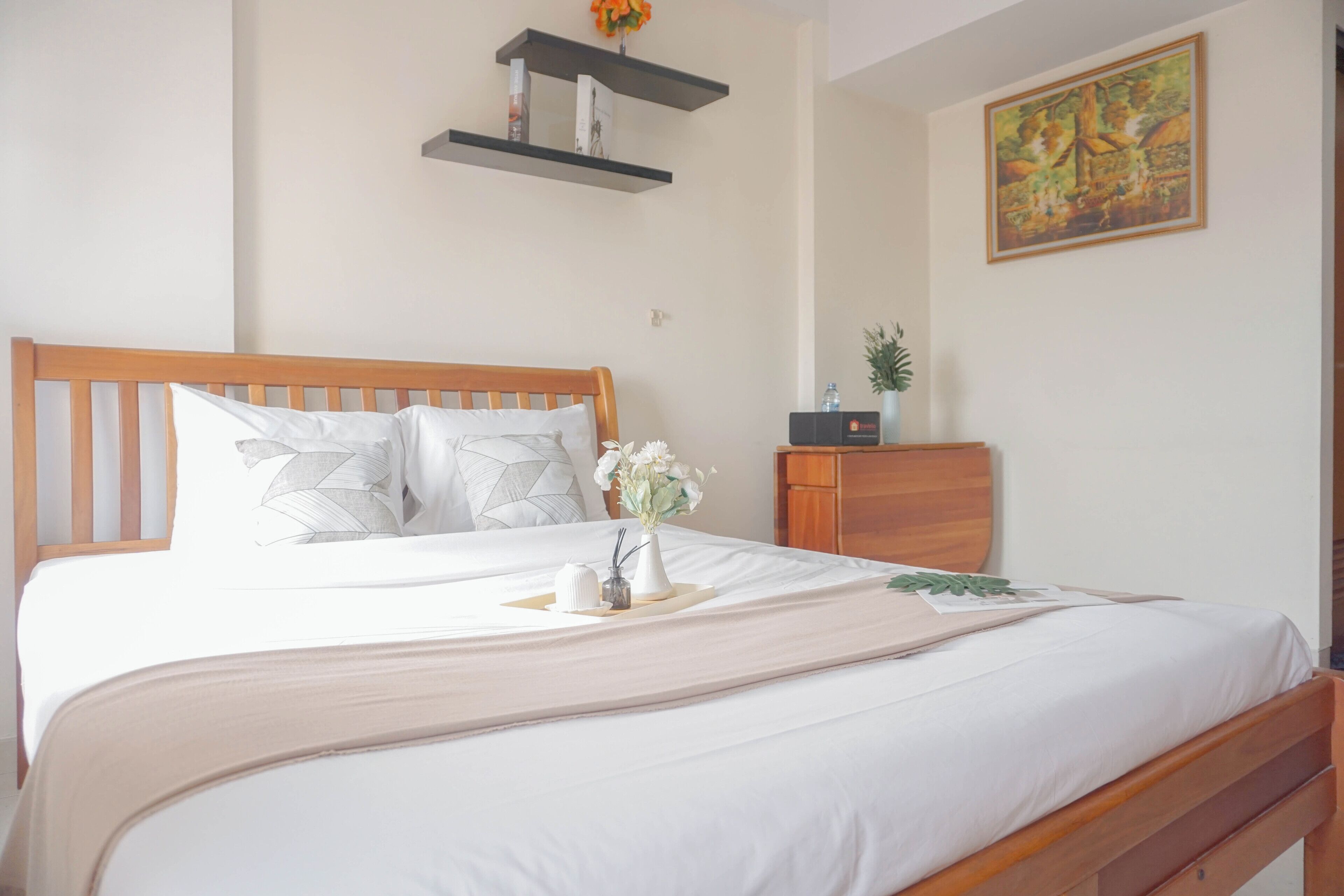 Una cama bien hecha con cabecera de madera, una mesita de noche con un florero y una estantería con libros, además de un cuadro en la pared.