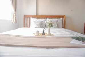 Een opgemaakt bed met een houten hoofdeinde, witte beddengoed en een dienblad met een bloemenvaas.