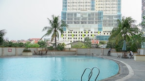 A swimming pool with a curved edge, surrounded by palm trees and a building with a modern design in the background. - Serene Studio Apartment At Margonda Residence 2 (Depok)