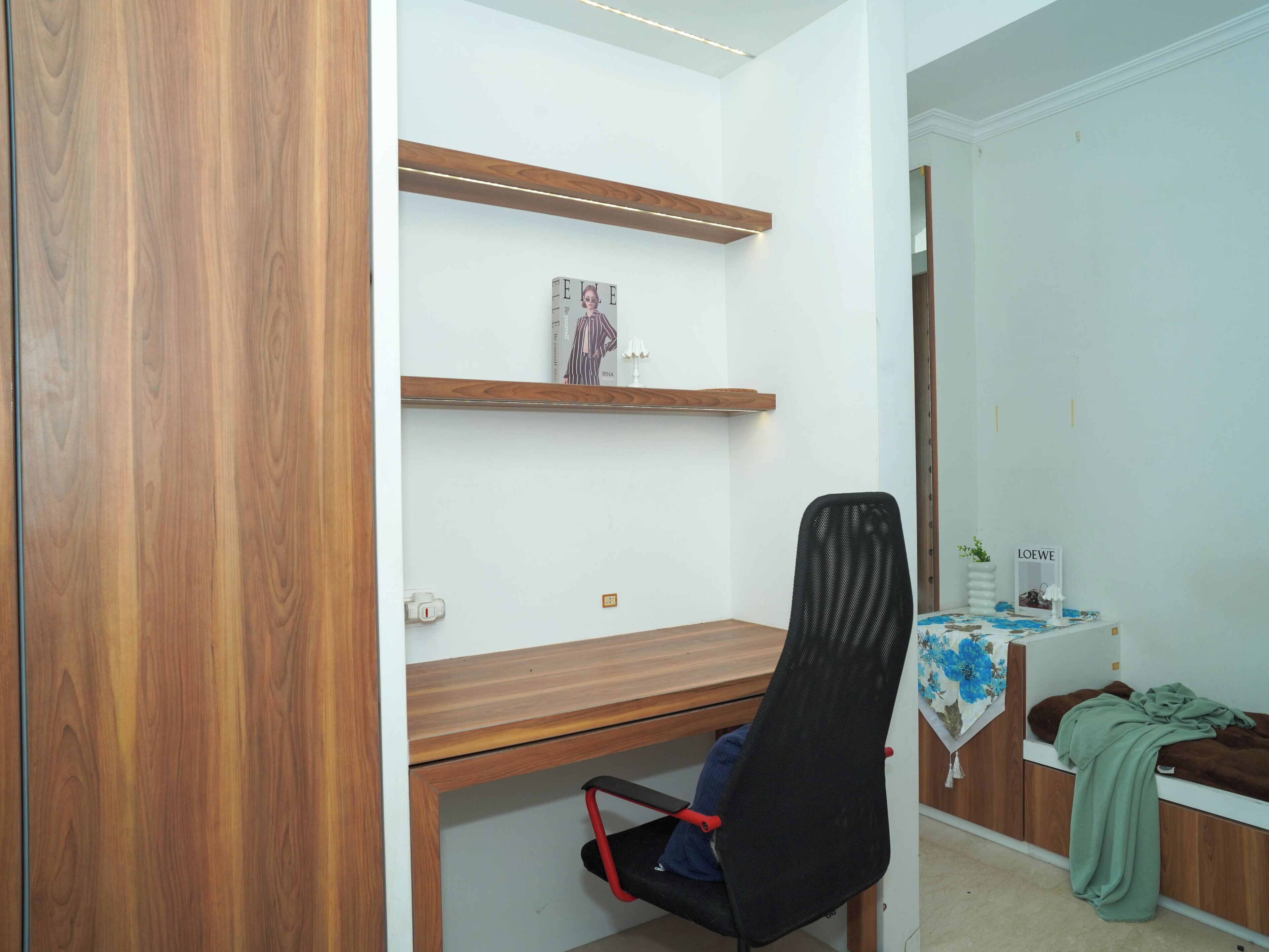 Ein Zimmer mit einem Holztisch, einem schwarzen Bürostuhl, einem kleinen Tisch mit einer blumigen Tischdecke und einem Spiegel mit dem Schriftzug 'LOEWE'.