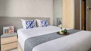 A neatly made bed with a grey blanket, two pillows with blue patterns, a bedside table with a book and a tray with flowers. - Wooden Accents Studio At Monroe Tower Apartment (Cikarang)